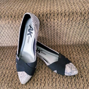 May include: A pair of pointed-toe flats featuring a grey and white snakeskin pattern. Each shoe has a black elastic cross-strap across the vamp. The interior insole is light grey and displays the black text AK SPORT and ANNE KLEIN.