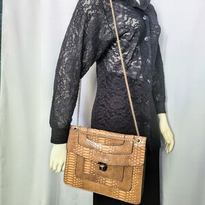 May include: A tan snakeskin-patterned handbag with a gold chain strap. The bag has a rectangular shape with a flap closure and a gold-tone clasp. The bag is displayed on a mannequin wearing a black lace top and black skirt.