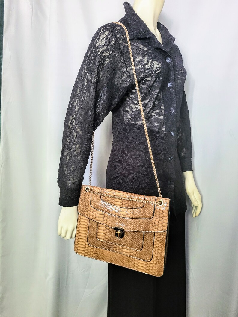 May include: A tan snakeskin-patterned handbag with a gold chain strap. The bag has a rectangular shape with a flap closure and a gold-tone clasp. The bag is displayed on a mannequin wearing a black lace top and black skirt.