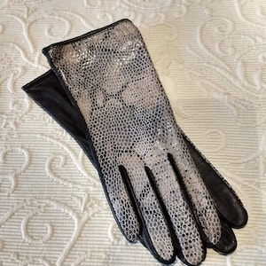 Könnte beinhalten: Ein Paar Handschuhe mit Schlangenmuster auf der Oberseite und schwarzem Leder an Handfläche und Fingern. Die Handschuhe liegen auf einer weißen, strukturierten Oberfläche.