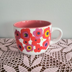May include: White ceramic mug with a pink interior and a handle. The mug is decorated with a repeating pattern of red, pink, and yellow flowers. The mug is sitting on a pink lace doily.