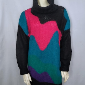 May include: A long-sleeved, oversized sweater dress with a black turtleneck and sleeves. The body of the dress features a vibrant, color-blocked design in shades of pink, teal, green, and purple. The dress is displayed on a mannequin.