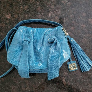 May include: A bright blue handbag with a snakeskin-like texture, featuring two long decorative fabric ties on the front, a matching shoulder strap, and a long blue tassel attached to a gold-toned metal charm with an intricate cutout design.