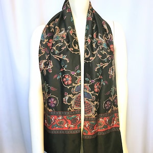 May include: Black scarf with a colorful floral and paisley pattern. The scarf has a red, teal, and gold border. The scarf is displayed on a mannequin.