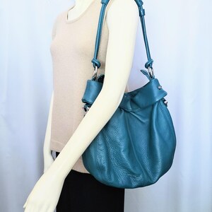 May include: Teal leather shoulder bag with a gathered design. The bag has a long strap and silver-tone hardware. The bag is displayed on a mannequin wearing a beige top and black skirt.