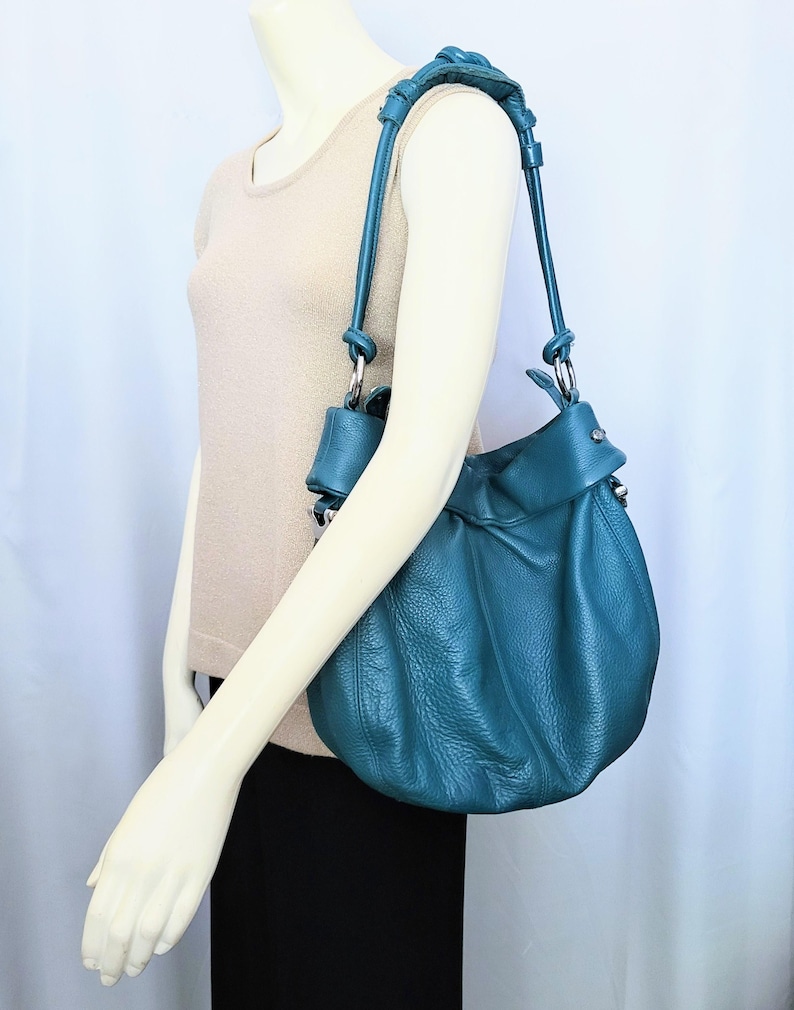 May include: Teal leather shoulder bag with a gathered design. The bag has a long strap and silver-tone hardware. The bag is displayed on a mannequin wearing a beige top and black skirt.