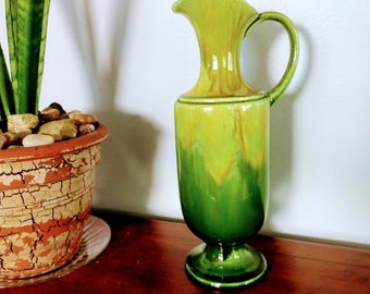 May include: A tall, green ceramic pitcher with a handle and a flared spout. The pitcher has a two-tone color scheme, transitioning from a lighter green at the top to a darker green at the base. It stands on a pedestal base.