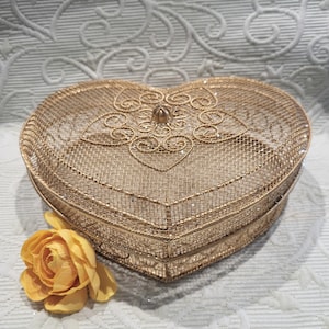 May include: A heart-shaped, gold-colored wire mesh box with a decorative filigree design on the lid. The box is embellished with small, sparkling accents. A yellow rose is placed near the box, adding a touch of color.