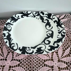 May include: Two white ceramic plates with a black floral scroll design around the rim. The plates are stacked on top of each other, showcasing the contrasting black and white color scheme. The plates are on a pink lace tablecloth.