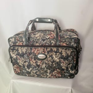 May include: A vintage travel bag with a floral tapestry pattern in shades of grey, brown, and pink. The bag features a top handle, a front zipped pocket, and a Pierre Cardin logo. The bag is designed for travel and has a classic, retro aesthetic.
