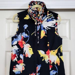 May include: A sleeveless vest with a navy blue background and a vibrant floral pattern in red, yellow, blue, and white. The vest has a full-length zipper and a stand-up collar. The vest is hanging on a beige hanger.
