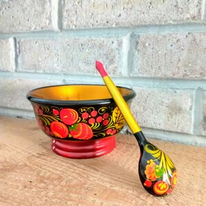 May include: A hand-painted wooden bowl and spoon set. The bowl is black with red, yellow, and green floral designs and a red base. The spoon is black with yellow accents and a red tip.