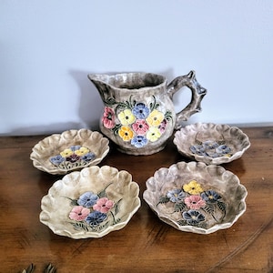 May include: A ceramic pitcher and four small dishes. The pitcher and dishes are beige with a floral design in pink, yellow, and blue. The pitcher has a textured surface and a decorative handle. The dishes have a scalloped edge.