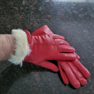 May include: A pair of bright red leather gloves featuring a soft, cream-colored faux fur trim at the wrist. The gloves have visible stitching details along the back of the hand and fingers, providing a structured look for cold weather.