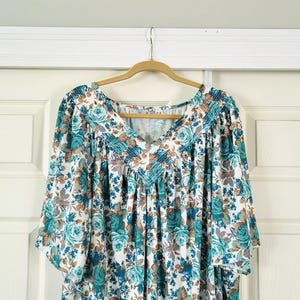May include: A vintage floral print dress with a V-neck and short sleeves, hanging on a wooden hanger. The dress features a white background with teal and brown rose patterns. The fabric appears soft and flowing.