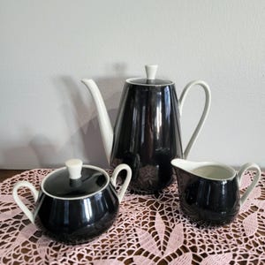 May include: A black and white tea set, including a teapot, sugar bowl, and creamer. The teapot has a tall, conical shape with a white handle and spout. The sugar bowl has a lid, and the creamer has a curved handle. The set is on a pink lace doily.