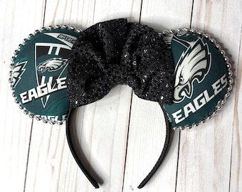 May include: A headband featuring two round ears covered in dark teal fabric with the Philadelphia Eagles logo and the word EAGLES printed in white. A large, black, glittery fabric bow sits between the ears, and the edges are lined with silver beads.