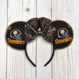 May include: Black and gold mouse ears headband featuring the Pittsburgh Steelers logo. The ears are black with a gold beaded trim. A sequined bow sits between the ears. The Steelers logo and text are in gold.