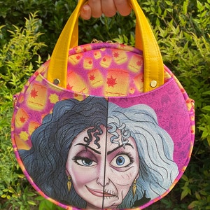 May include: A round pink and yellow purse with a yellow handle. The front of the purse features a colorful illustration of the character Maleficent from the Disney movie Sleeping Beauty. The purse has a pink background with yellow lanterns and a yellow background with pink lanterns. The text "Kingdom Ears" is printed in yellow at the bottom of the image.