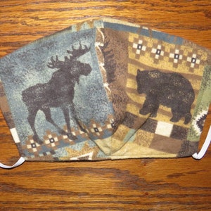 May include: A brown and blue fabric face mask with a moose and bear design. The mask has white elastic straps.