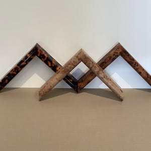 Puede incluir: Tres esquinas de marcos de fotos de madera dispuestas en forma de V. Los marcos tienen diferentes acabados: uno con un patrón de carey, otro con veta de madera clara y otro con veta de madera más oscura. Los marcos están colocados sobre una superficie neutra.