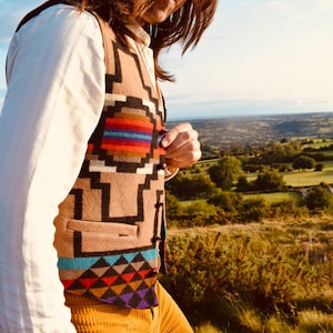 May include: A tan waistcoat featuring a geometric design in black, red, blue, and purple. It's worn over a white shirt and paired with mustard corduroy trousers. The background shows a scenic outdoor view.