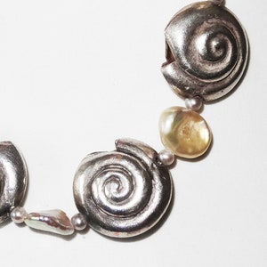 Op de afbeelding: Zilverkleurige ketting met spiraalvormige hangers en onregelmatige parels. De ketting combineert metalen en natuurlijke elementen. De parels zijn in crème- en goudtinten. Klassiek ontwerp.