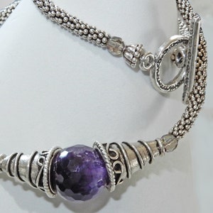 Op de afbeelding: Zilverkleurige ketting met een grote, facetgeslepen paarse edelsteen als middelpunt. De ketting heeft een getextureerd, kralenkettingontwerp en decoratief metaalwerk rond de edelsteen. De sluiting is een tuimelsluiting.
