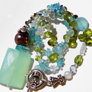 May include: A multi-strand necklace featuring a large, light green rectangular pendant and a dark brown, iridescent bead. The necklace incorporates various shades of blue and green gemstones, including faceted and irregular shapes, with a silver-toned clasp.