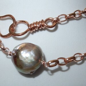 May include: A copper-toned necklace featuring a large, iridescent pearl pendant. The necklace has a chain link design with a unique hook clasp. The pearl is a mix of silver, brown, and green.