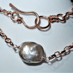 May include: A copper-colored necklace featuring a large, irregularly shaped pearl pendant. The necklace has a chain link design with a matching clasp. The pearl has a lustrous, iridescent surface, reflecting shades of white, gray, and pink.