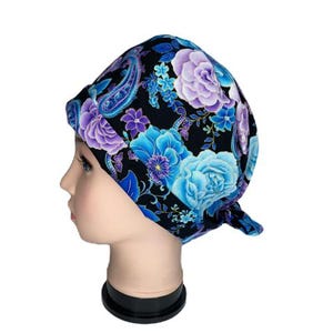 May include: A scrub cap featuring a black background with a vibrant floral and paisley pattern in shades of light blue, turquoise, and purple. The fabric head covering has a rounded crown and a tie-back closure, displaying large blooming flowers.