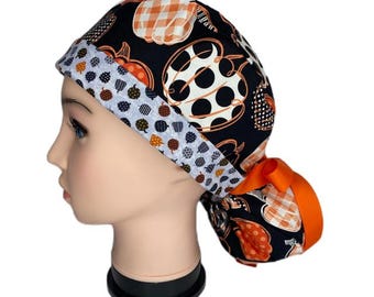 Pode incluir: Uma touca cirúrgica com estampa preta e laranja com abóboras e uma borda de bolinhas brancas e cinzas. O chapéu tem uma fita laranja e foi projetado para ser usado durante a estação do outono. O chapéu está em uma cabeça de manequim.