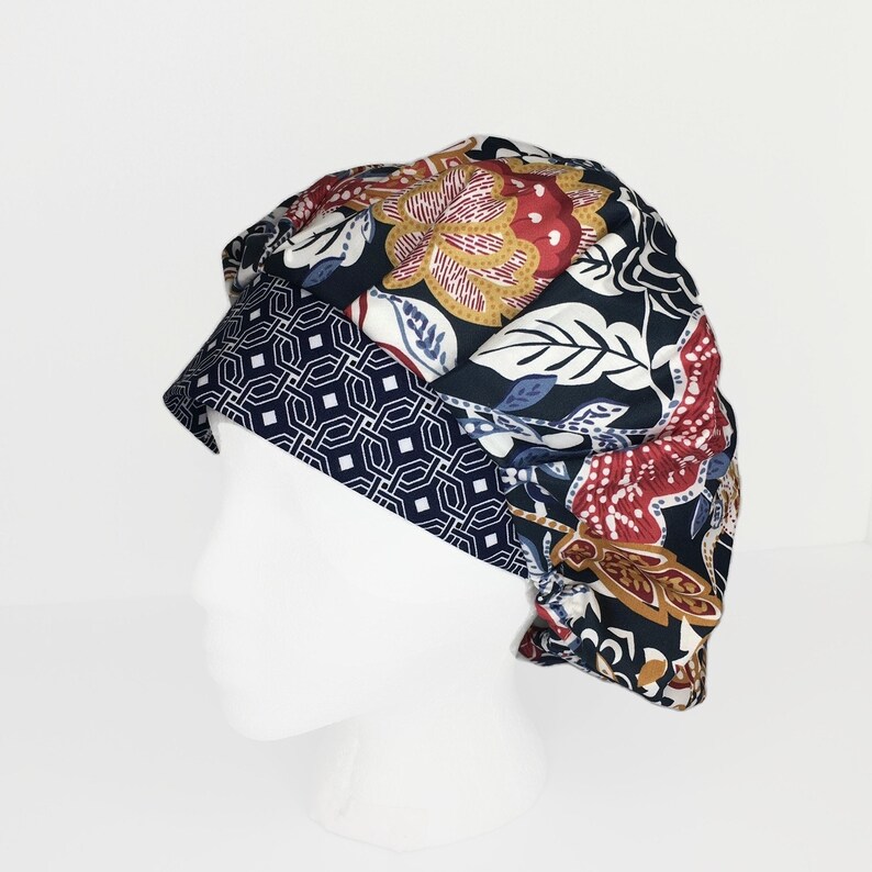 Puede incluir: Un gorro quir&uacute;rgico estampado con un fondo azul marino oscuro y un dise&ntilde;o floral en rojo, dorado y blanco. El borde presenta un patr&oacute;n geom&eacute;trico en azul marino y blanco. El gorro se muestra en una cabeza de maniqu&iacute; blanca.