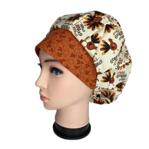 May include: A Thanksgiving-themed scrub hat featuring a cream-colored background with brown turkeys and the words "Gobble Gobble." The brim is a solid orange with a floral pattern. The hat is designed to be worn during medical procedures.