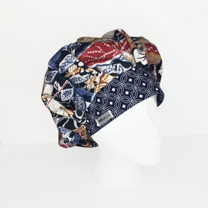 Puede incluir: Un gorro de exfoliaci&oacute;n abullonado estampado con interior blanco. El gorro presenta una mezcla de patrones florales y geom&eacute;tricos en azul marino, rojo y dorado. Una etiqueta blanca con texto es visible en la parte delantera. El gorro se muestra en una cabeza de maniqu&iacute; blanca.