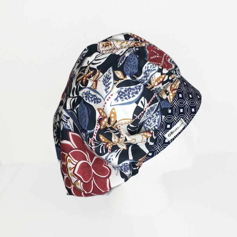 Puede incluir: Un gorro quir&uacute;rgico estampado con un dise&ntilde;o floral en tonos azul marino, burdeos, blanco y dorado. El gorro tiene una banda con estampado geom&eacute;trico y el texto "ORhats.com" en el lateral.
