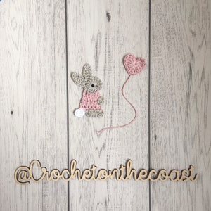 May include: A crocheted bunny holding a pink heart balloon. The bunny is wearing a pink dress and has a white tail. The background is a white wooden surface. The text "@CrochetonTheCoast" is written on the surface.