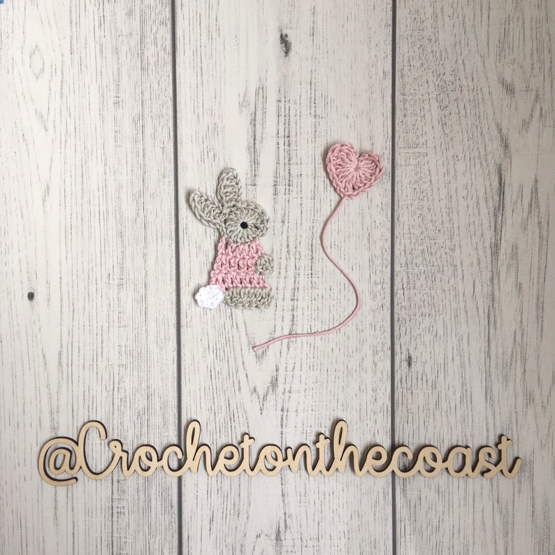 May include: A crocheted bunny holding a pink heart balloon. The bunny is wearing a pink dress and has a white tail. The background is a white wooden surface. The text "@CrochetonTheCoast" is written on the surface.