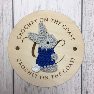 May include: A crocheted gray bunny wearing a blue crocheted sweater. The bunny is attached to a wooden round sign with the text "Crochet on the Coast" written on it.