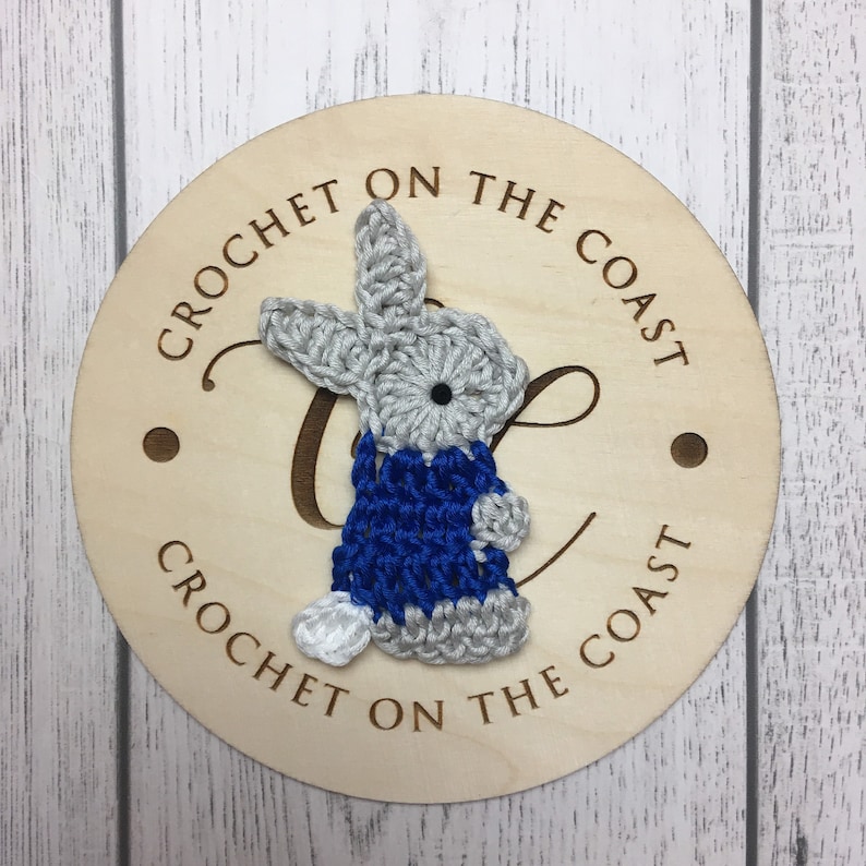 May include: A crocheted gray bunny wearing a blue crocheted sweater. The bunny is attached to a wooden round sign with the text "Crochet on the Coast" written on it.