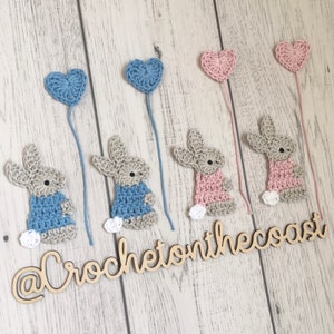 May include: Five crocheted bunny ornaments with heart-shaped balloons attached. The bunnies are in blue, pink, and beige. The balloons are in blue and pink. The ornaments are hanging from a wooden surface with the text "@CrochetonTheCoast" written on it.