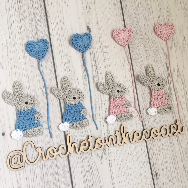 May include: Five crocheted bunny ornaments with heart-shaped balloons attached. The bunnies are in blue, pink, and beige. The balloons are in blue and pink. The ornaments are hanging from a wooden surface with the text "@CrochetonTheCoast" written on it.