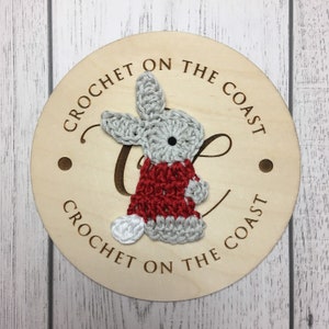May include: A wooden round sign with the text "Crochet on the Coast" written around the edge. A crocheted gray bunny wearing a red sweater is attached to the sign.