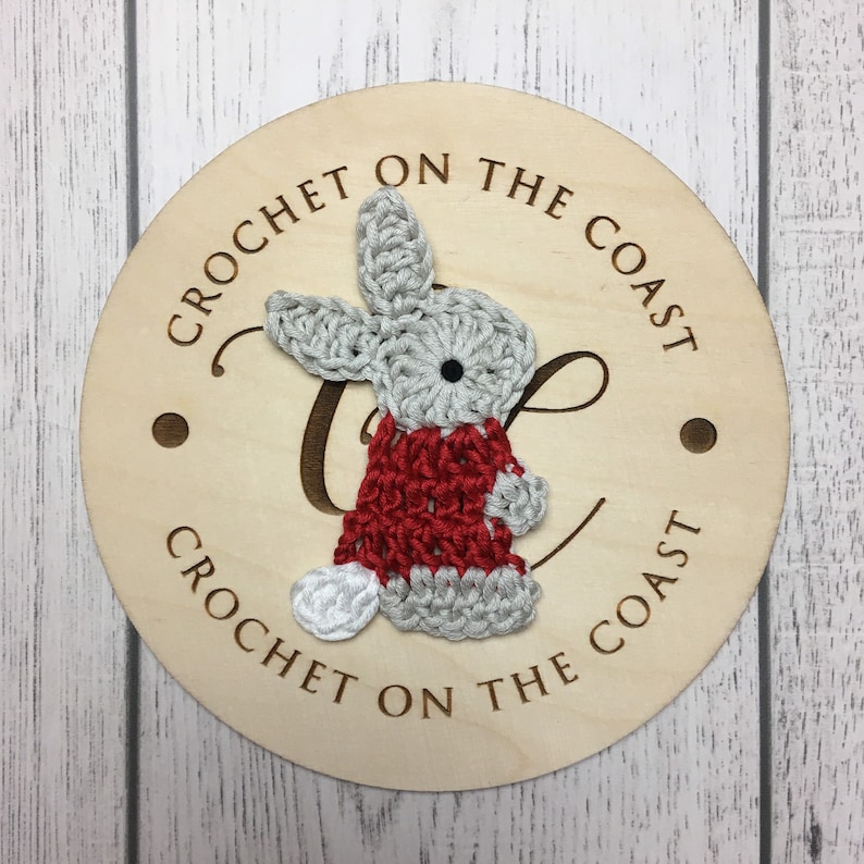May include: A wooden round sign with the text "Crochet on the Coast" written around the edge. A crocheted gray bunny wearing a red sweater is attached to the sign.
