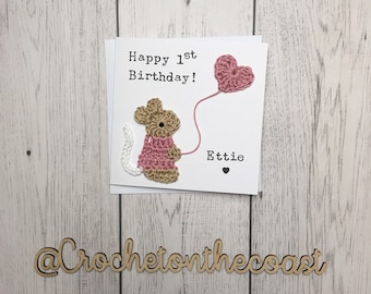 May include: A handmade crochet card with a pink mouse holding a pink heart balloon. The card says "Happy 1st Birthday!" and "Ettie" with a small heart. The card is on a wooden background with the text "@CrochetonTheCoast" in a wooden font.