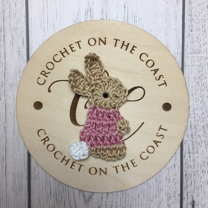 May include: A wooden round sign with the text "Crochet on the Coast" written around the edge. A crocheted bunny in pink and beige is attached to the sign.