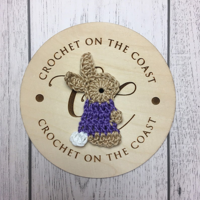 May include: Crocheted bunny wearing a purple dress on a wooden round sign with the text "Crochet on the Coast".