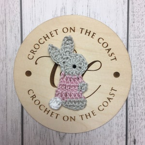May include: A crocheted bunny with a pink dress and gray ears, on a wooden round sign with the text "Crochet on the Coast" repeated.