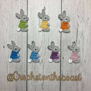 May include: A set of ten crocheted bunny appliques in various colors, including green, yellow, orange, blue, purple, and pink. The bunnies are all the same size and have gray ears and faces. The appliques are arranged in two rows of five on a white wooden background. The text "@CrochetonTheCoast" is written in brown on a wooden sign below the bunnies.